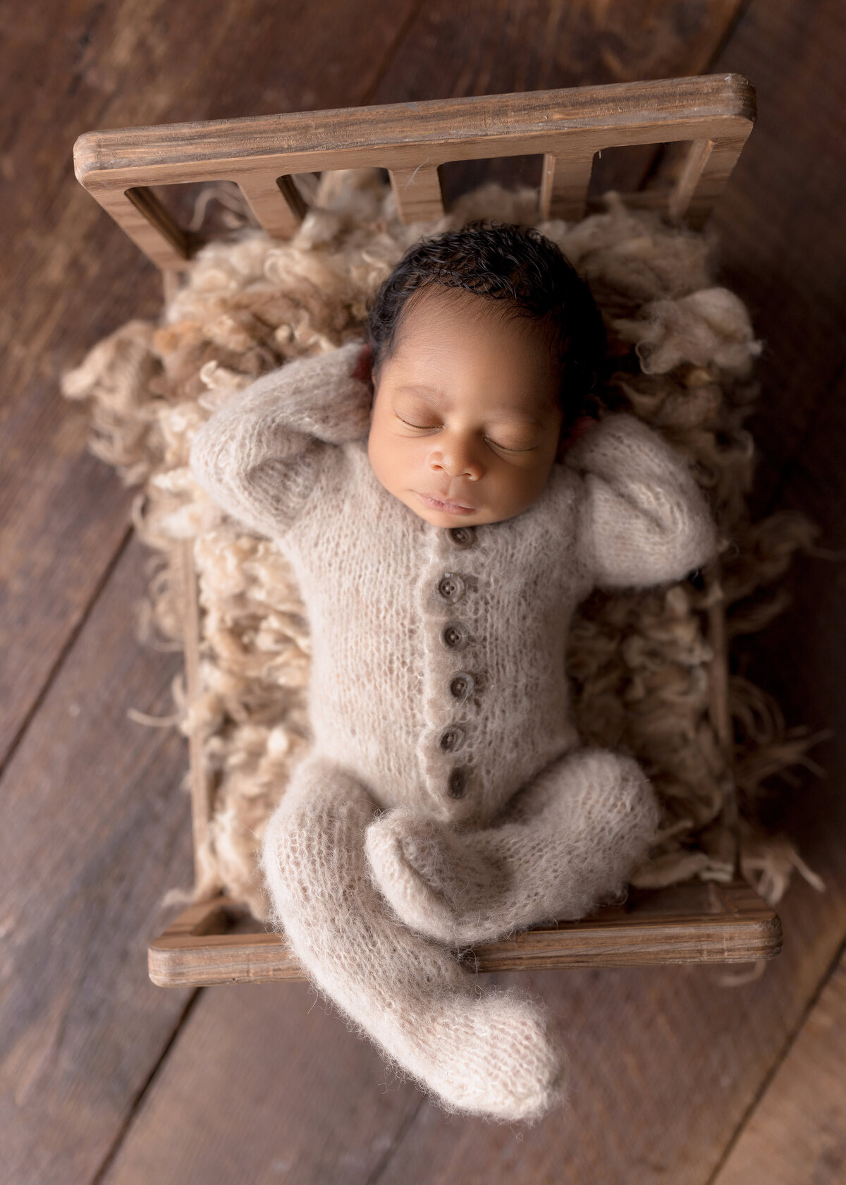 Newborn photoshoot in West Palm Beach Florida. Black baby is wearing a one piece cream-colored knit one-piece outfit laying on his back on a newborn bed prop. Aerial image. Baby's hands are resting under his head and his legs are sprawled atop of the foot of the miniature bed.
