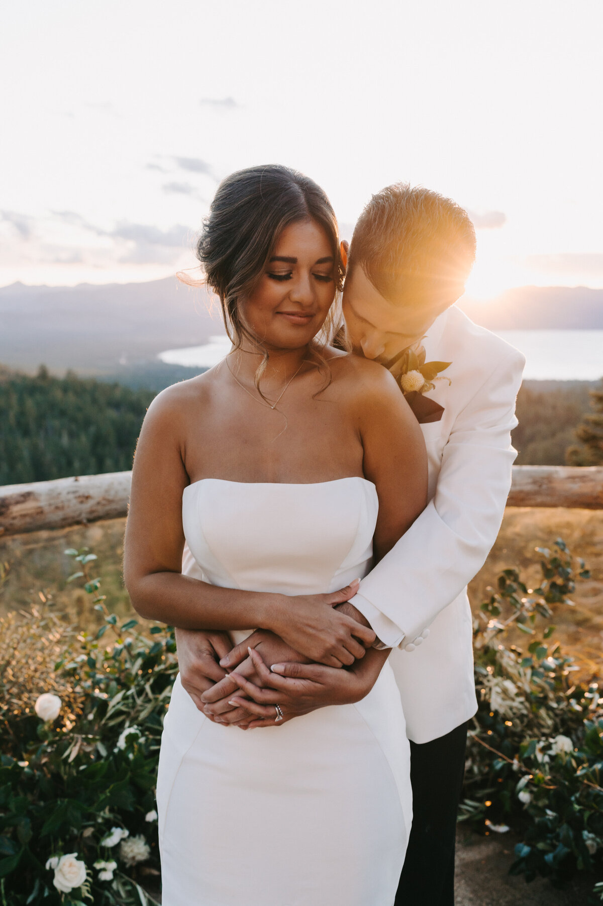 A colorful portrait of Michelle and Julien on their wedding day at Tahoe Blue Estate in Lake Tahoe, Nevada. The portrait is at sunset with the sun glowing behind the groom’s shoulder. The bride’s back is against the groom who has his arms wrapped around her waist while he’s kissing her on the shoulder. Her head is turned slightly towards him, her eyes downcast and she has a slight smile. The view of the lake and mountains are behind them. Wedding photographs by Stacie McChesney of Vitae Weddings.