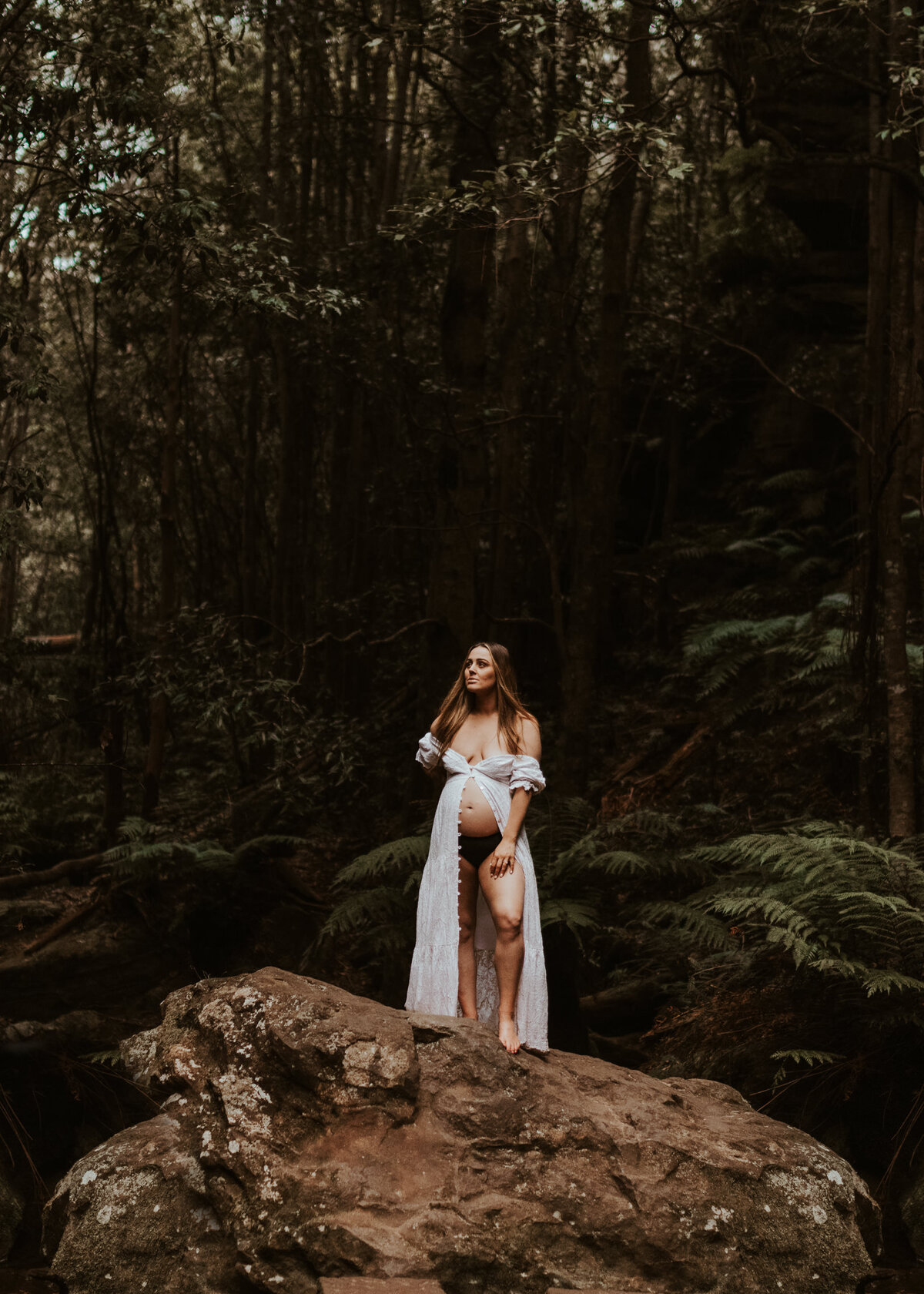 Pregnant woman in a white robe standing barefoot on a rock in a dense forest during a moody outdoor maternity session in Sydney