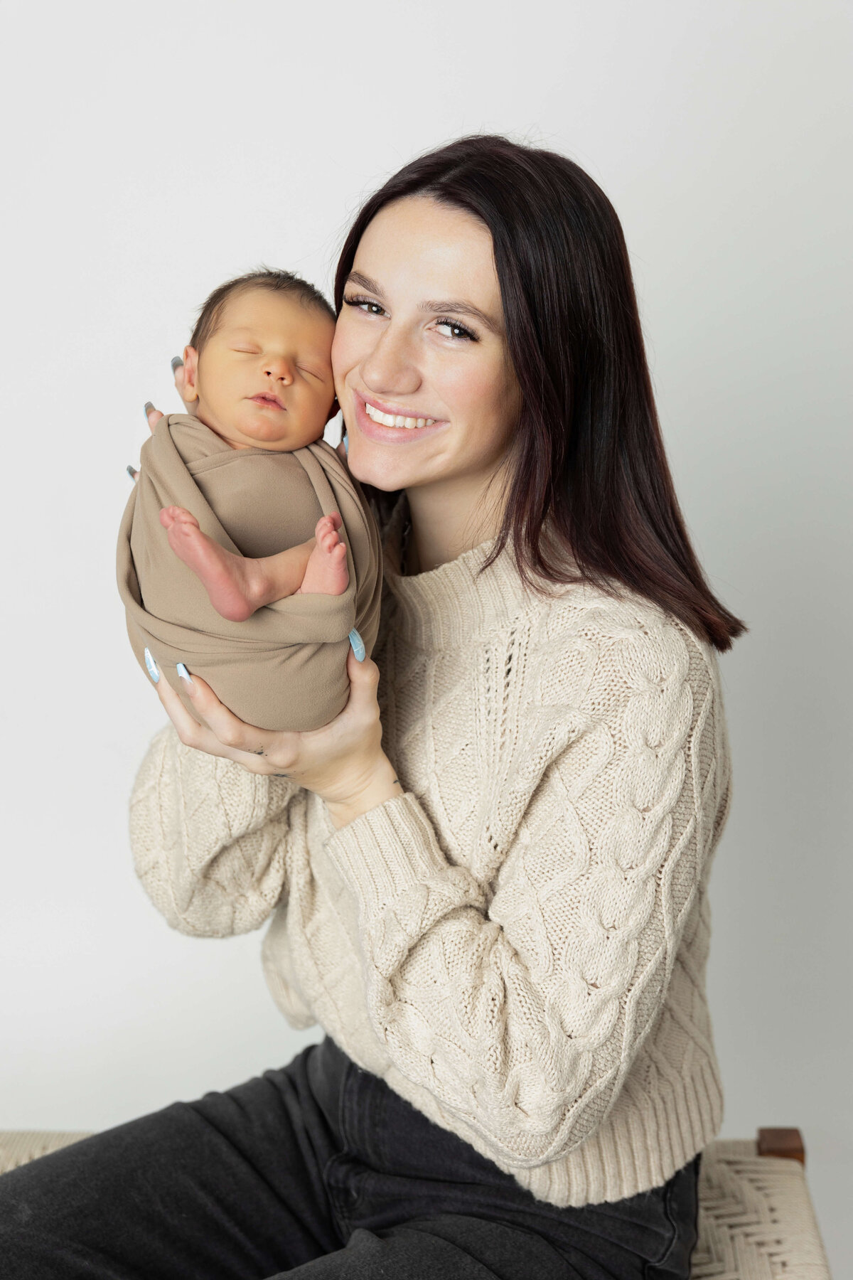 A joyful new mother smiles at the camera while holding her swaddled newborn in a soft brown wrap. They are seated against a plain, light-colored background that emphasizes their connection.