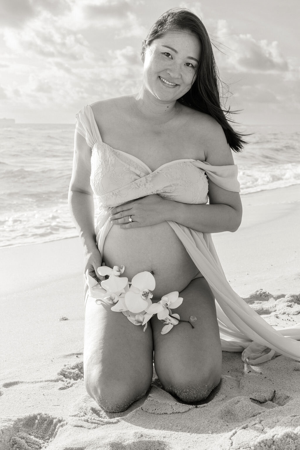 Maternity portrait of a woman kneeling on the sand with orchids during a sunrise beach session in Miami, South Florida.