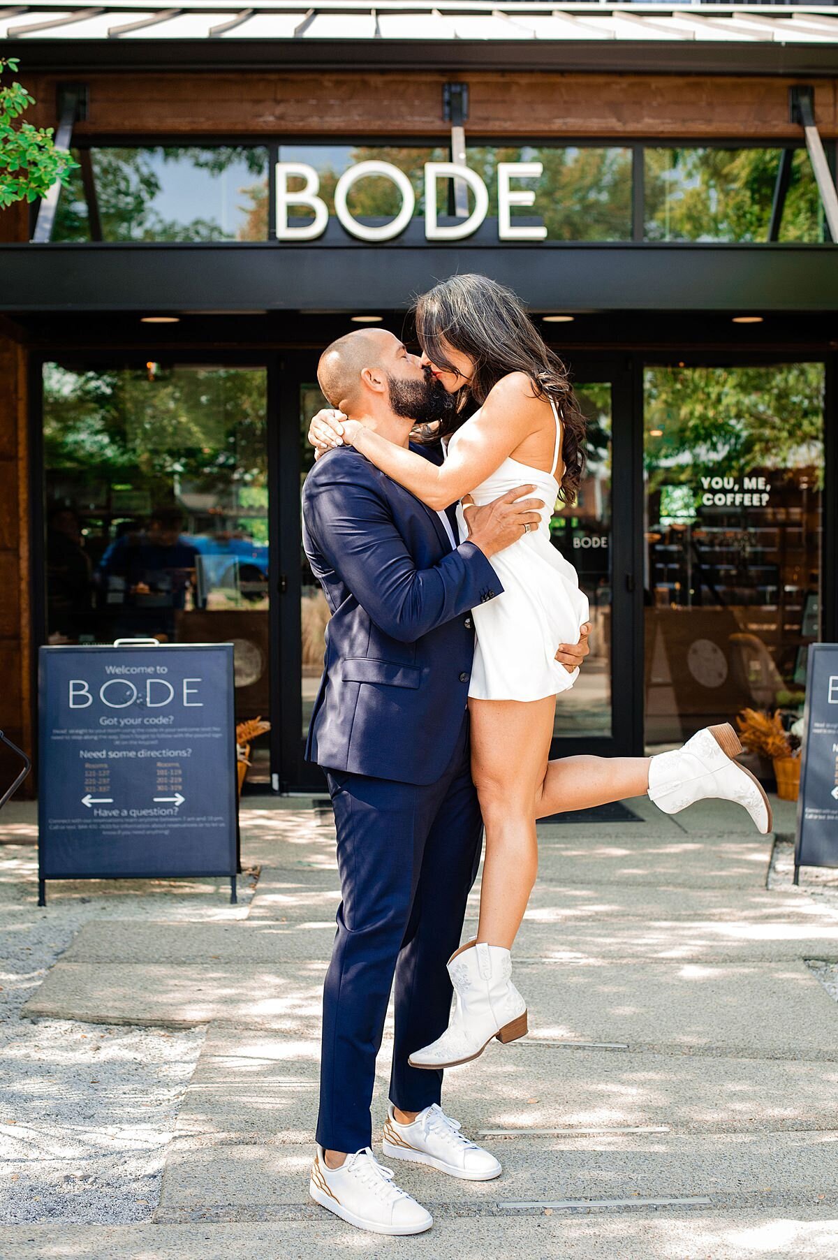 The groom, wearing a slim fit dark blue suit and white sneakers lifts up the bride as he kisses her in front of Bode Nashville. The bride is wearing a mini dress with white booties. She has her leg kicked up in the air as she kisses the groom.