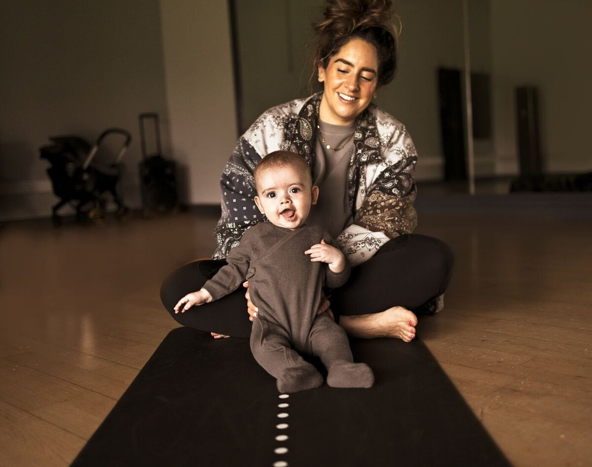A smiling mother sits cross-legged on a yoga mat with her young baby, who is looking at the camera with their tongue out, in a room in UK