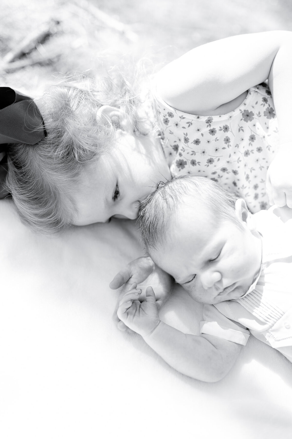 Newborn, sibling, outdoor, soft light, white blanket – A smiling young girl with a blue bow lies on a white blanket beside her newborn sibling dressed in white.