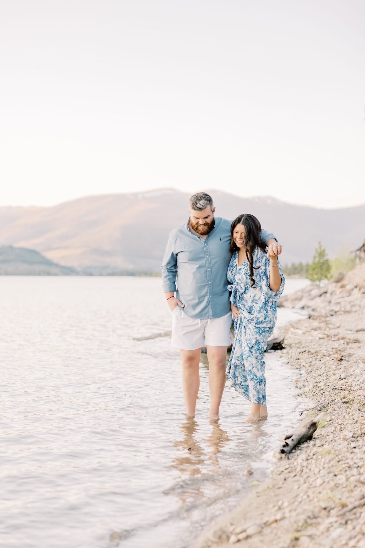 A man and woman are walking arm in arm. They are walking with their toes dipped in the water and are laughing with one another. 