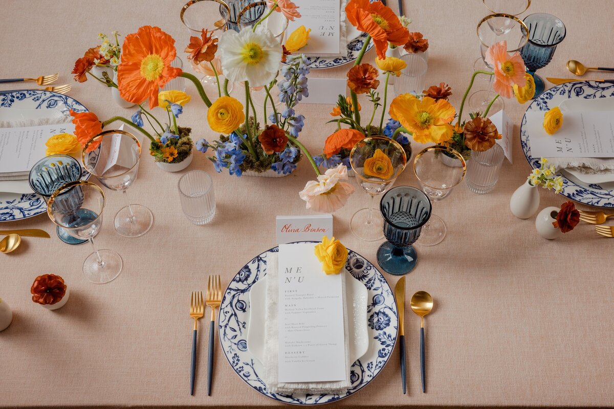 top-down view of a stylish wedding table featuring bright poppies, gold-rimmed plates, and detailed menu cards on a soft linen tablecloth
