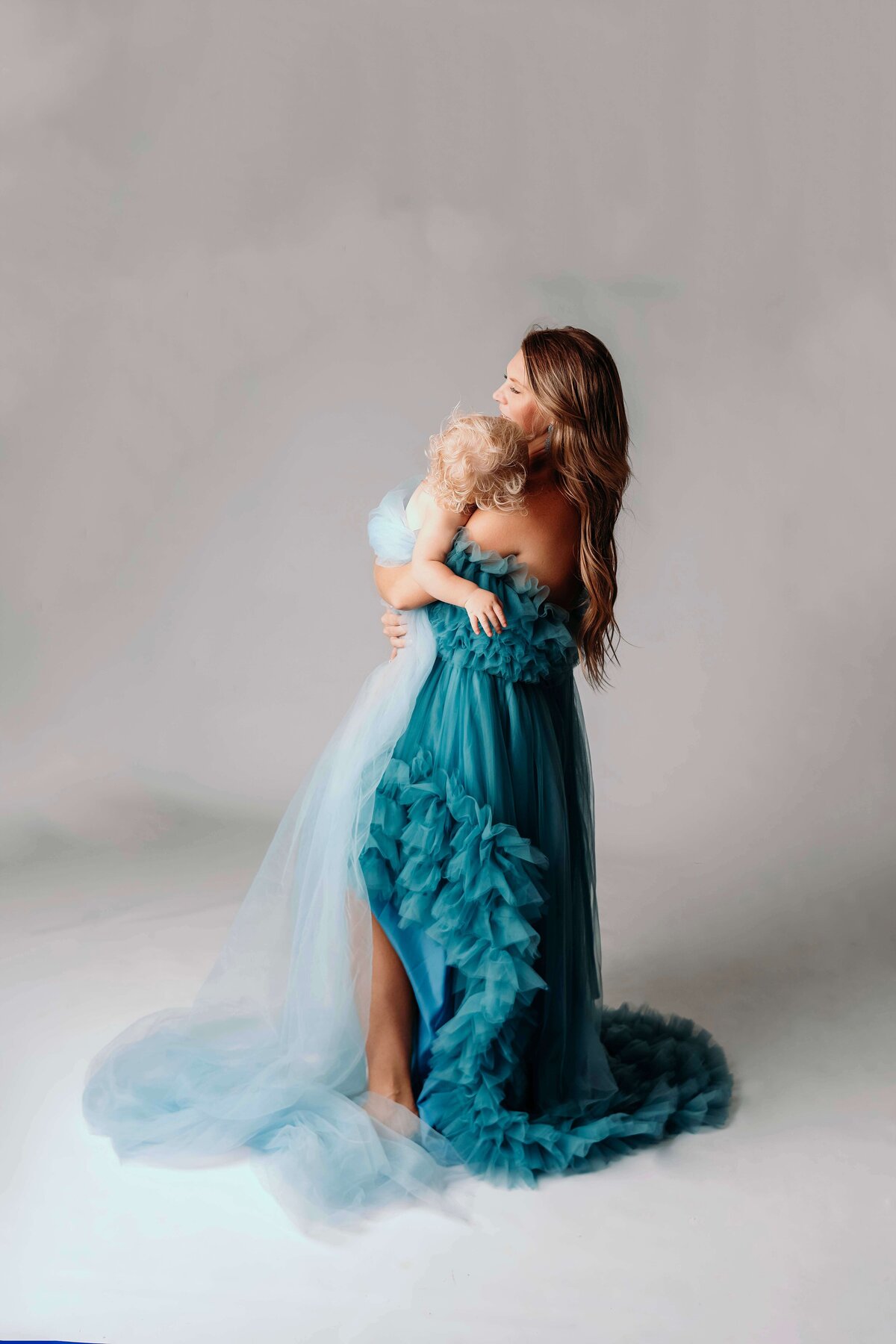 A young mom stands, gently holding her toddler daughter as the child rests her head on her shoulder. They are dressed in matching blue tulle gowns, with the little girl wearing a white floral headband. This tender moment is captured by Mobile, AL Motherhood Photographer, highlighting the closeness between them