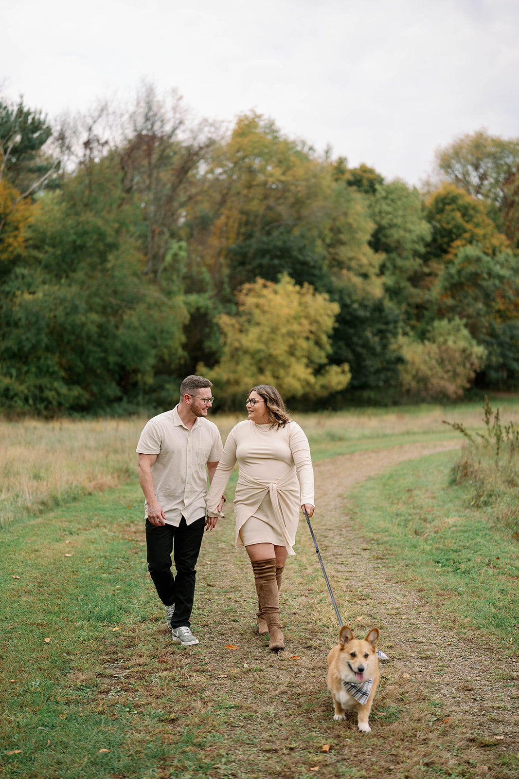 Couple walking hand in hand with their dog along a wooded trail during their engagement session at Al Sabo Preserve in Kalamazoo, Michigan.