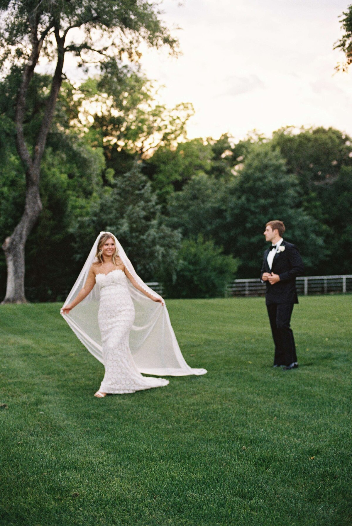 Bride in a strapless lace gown twirls with her veil on a grassy lawn while the groom in a black tuxedo looks on at sunset.