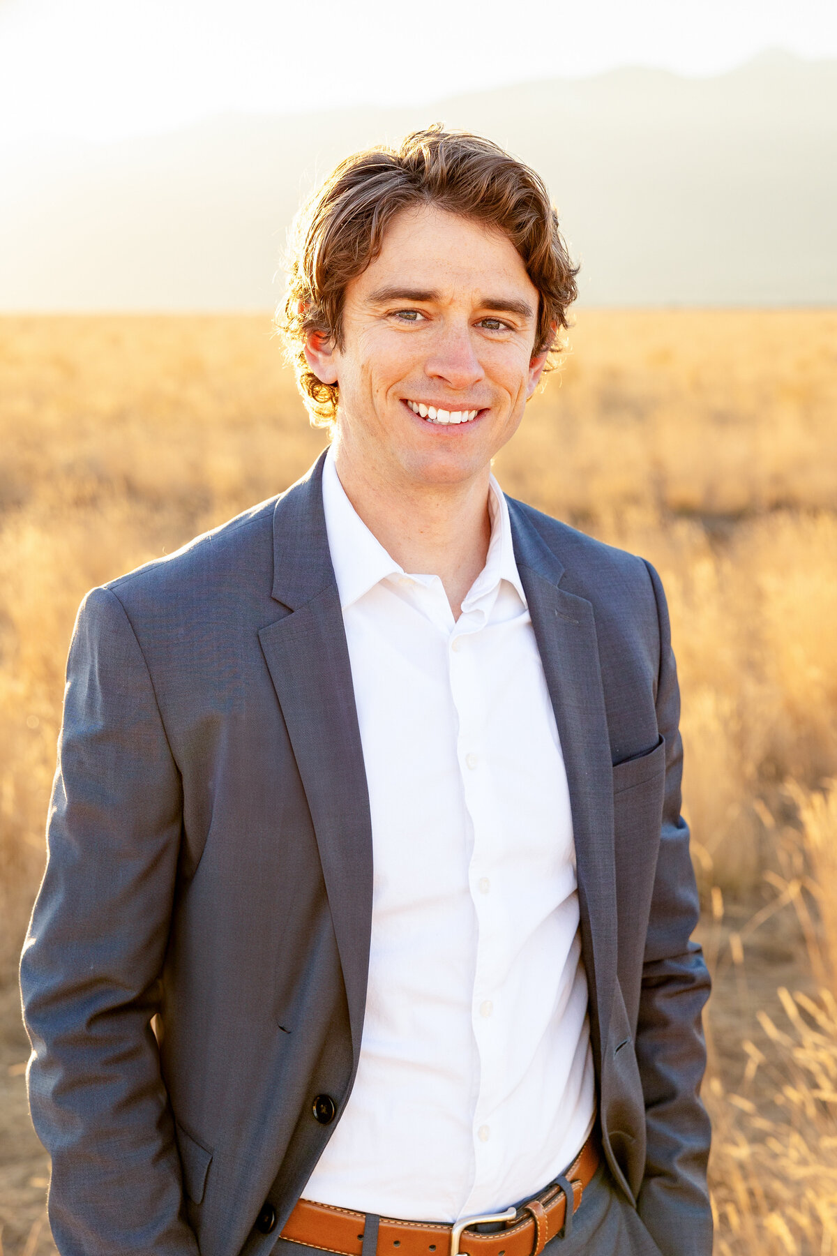 A father dressed in a navy suit and white shirt smiles confidently in a golden field, the sun setting behind him.