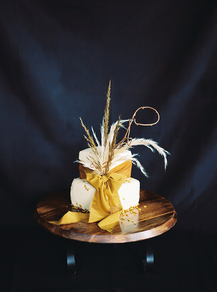 A white wedding cake with yellow ribbon and natural tan pampas on a wooden cake stand with a black backdrop