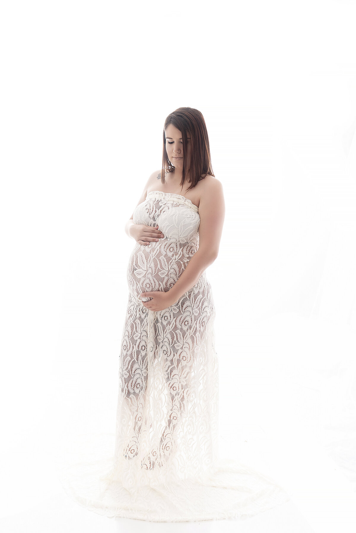 A mother to be wearing a lace see through maternity coverup stands in a studio holding her bump