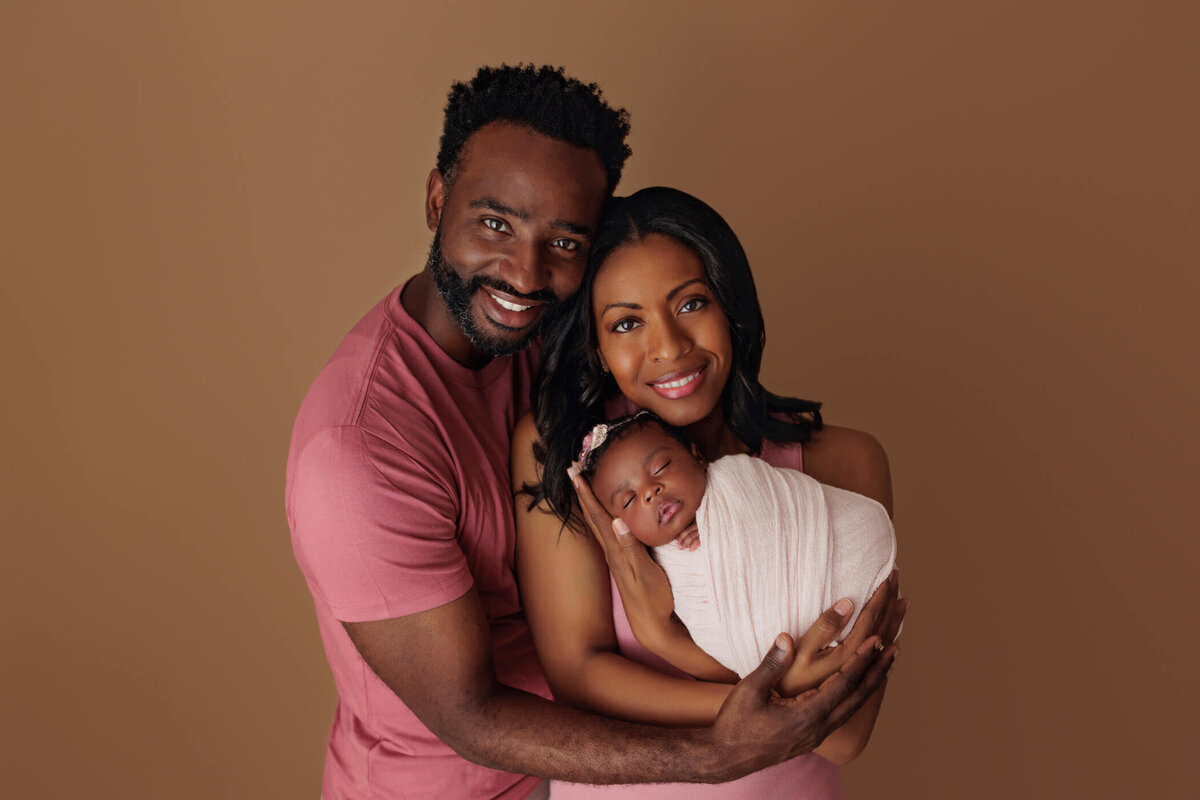 A happy couple, the father in a pink shirt and the mother in a pink dress, lovingly holding their newborn baby swaddled in white against a brown background.