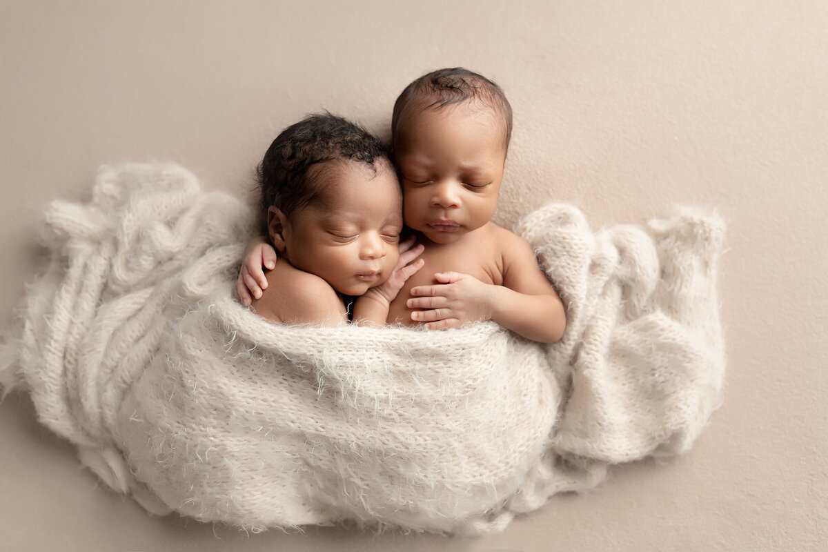 Newborn twin photoshoot in West Palm Beach and Jupiter photography studio. Twin Black baby boys are cuddled on a stretch fabric. One twin is laying on his back, with his arm wrapped around his brother's back. The other twin is sleeping on his brother's shoulder with his hand on his chest and resting his cheek on his hand.  The boys are draped in a cream colored knit swaddle.Aerial image.