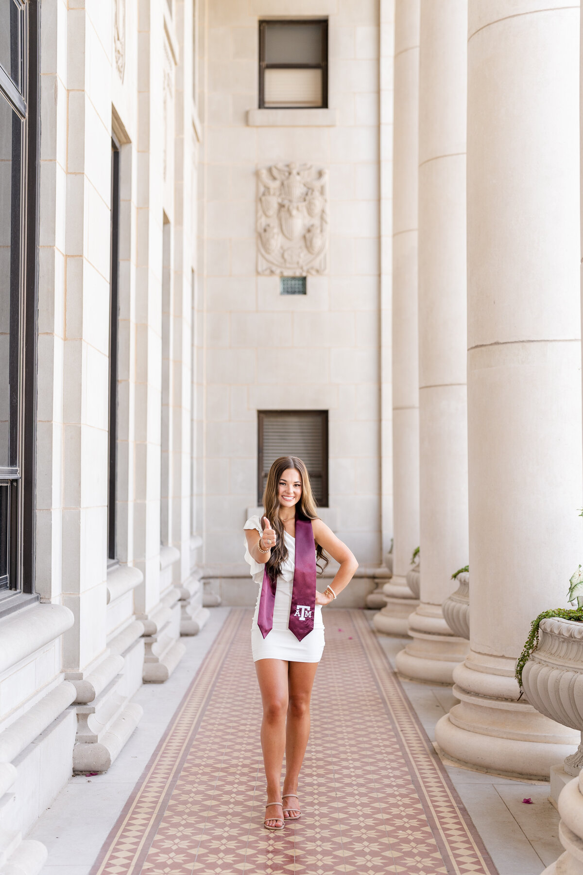 Texas A&M senior girl standing with hand on hip and thumbs up while wearing white dress and maroon stole in the columns of the Administration Building