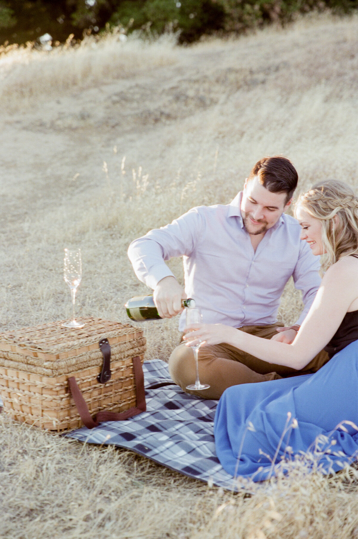 A man and her lady sit on a picnic rug at a meadow and celebrate their new engagement with bubble champagne from a wooden basket.