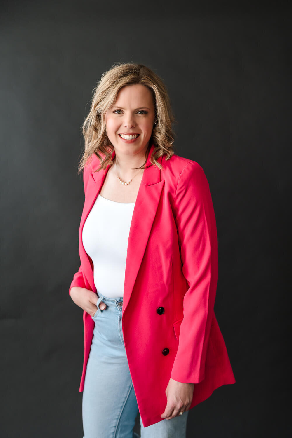 Smiling female entrepreneur wearing bright pink blazer and light jeans in Kelowna photo studio.