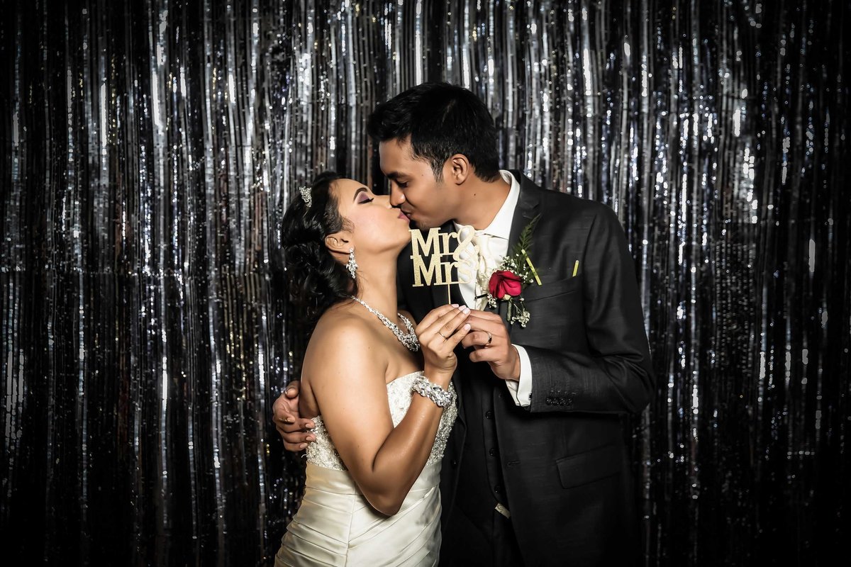 Bride and groom kiss in front of silver backdrop holding a "Mr & Mrs" sign. Photobooth by Ross Photography, Trinidad, W.I..