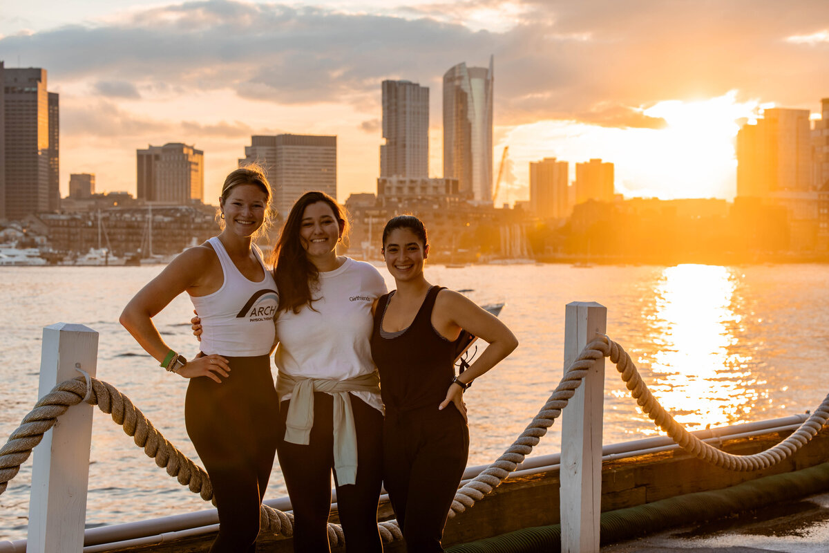 3 women standing in front of boston skyline during sunset.