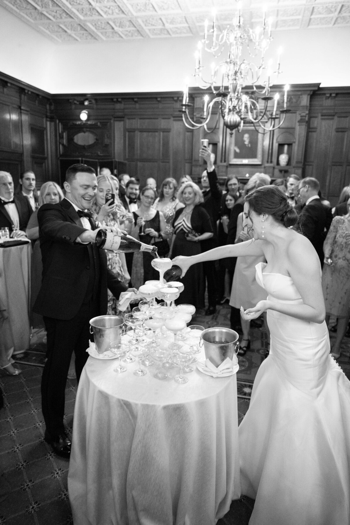 A bride and groom pour champagne into a tower of glasses at a crowded wedding reception. Guests, dressed in formal attire, enthusiastically watch and raise their glasses. The setting has elegant wood paneling and a chandelier overhead.