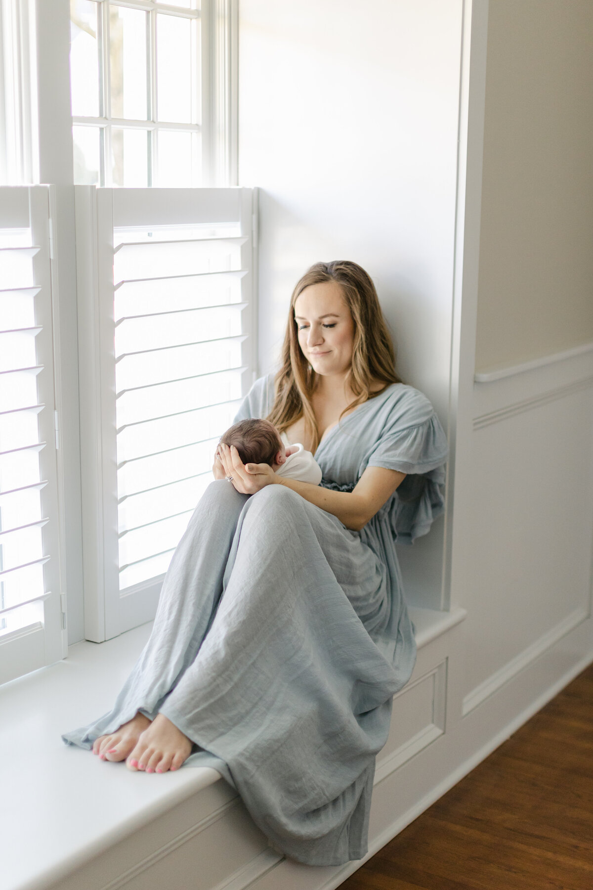 Mom wearing an elegant dusty blue dress sitting within a window holding her newborn baby boy photographed by Philadelphia Newborn Photographer Tara Federico