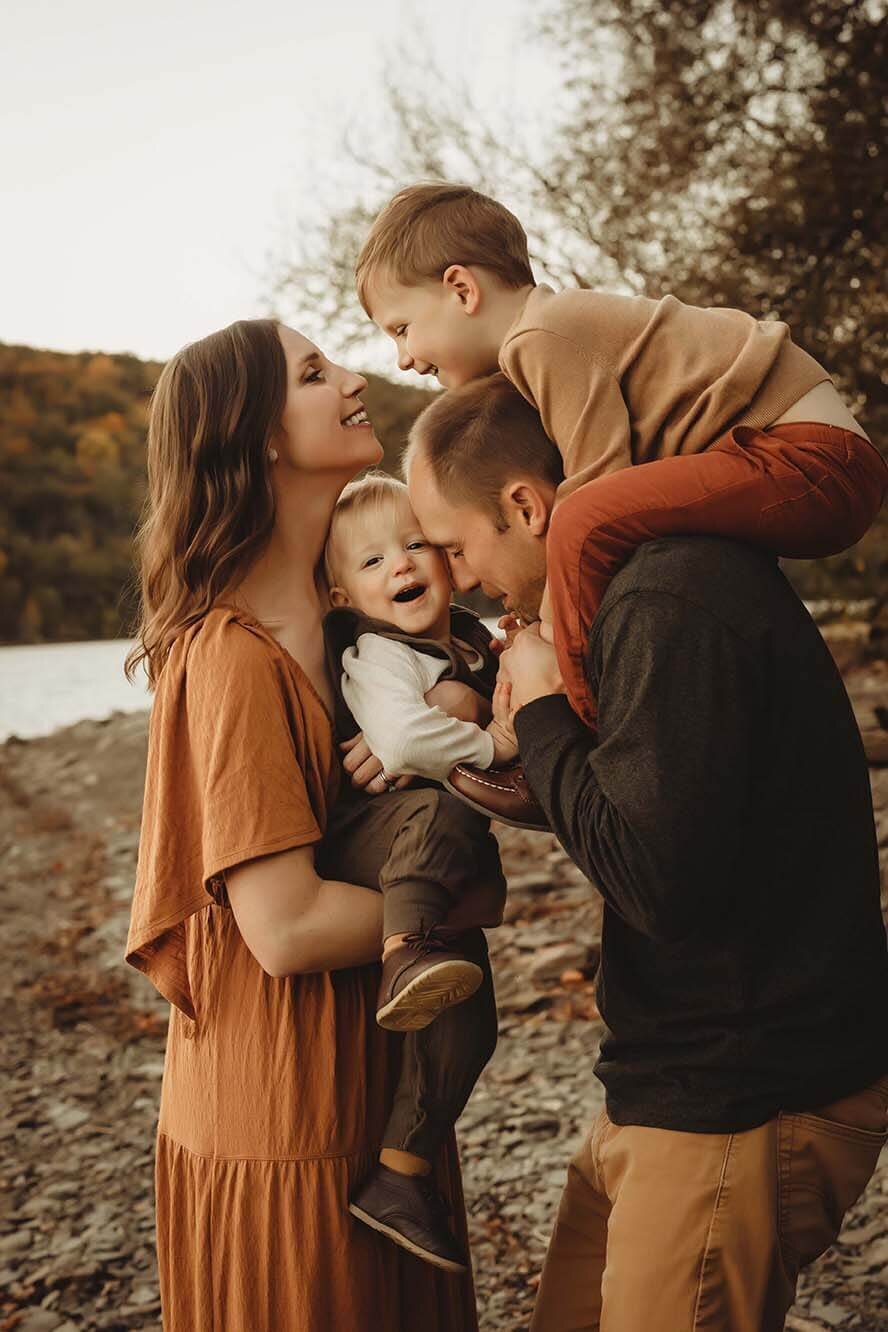A mom holds her youngest son to the dads face. Dad has his oldest son on his shoulders while the oldest son and the mom share a beautiful moment  on connection
