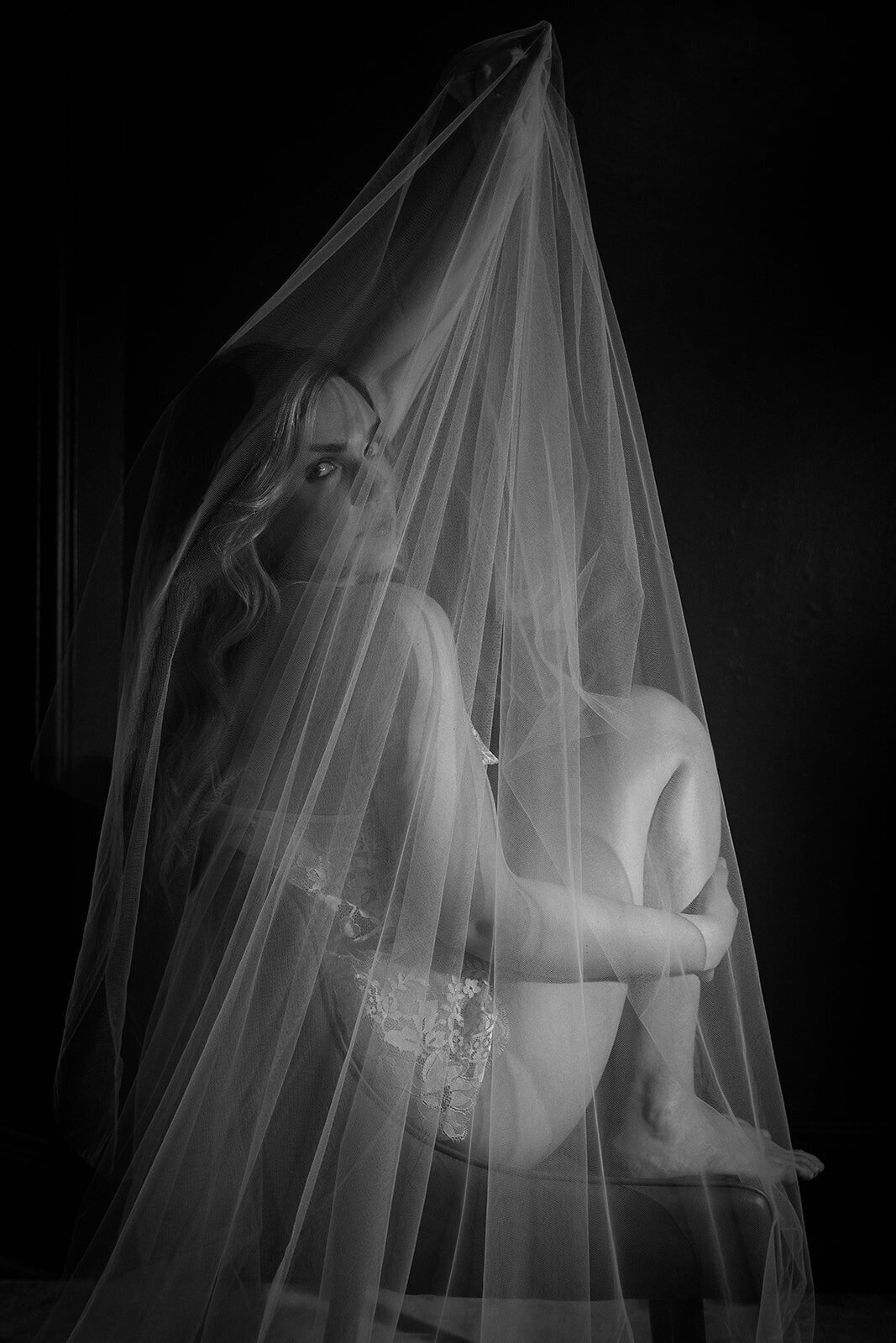 Intimate boudoir photo shoot bride sits in a chair holding her veil above her head as it drapes over her.