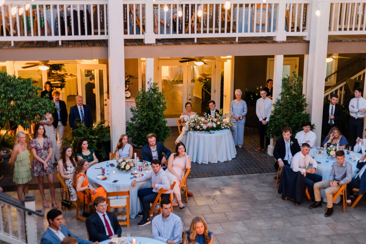 A wedding reception with guests seated at round tables outdoors, some standing and watching. A couple sits at a decorated table with string lights above.