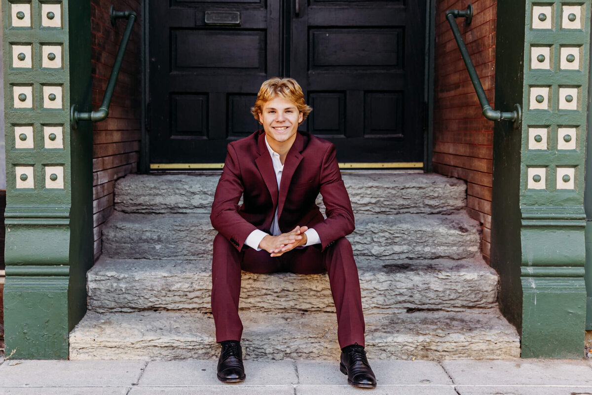 handsome blonde teenage boy in a burgundy suit sitting on stone steps and smiling captured by Ashley Kalbus Photography