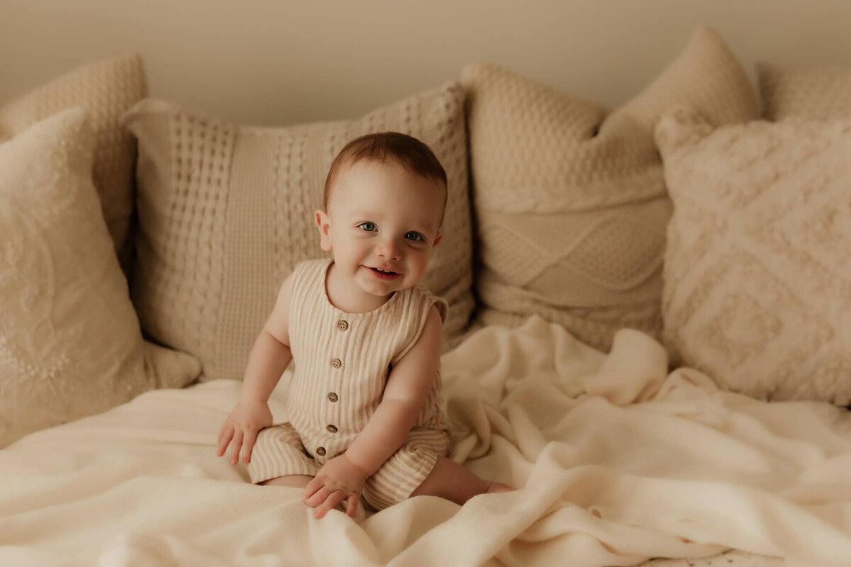 Little boy with red hair smiling while sitting on a bed wearing a beige romper with buttons.
