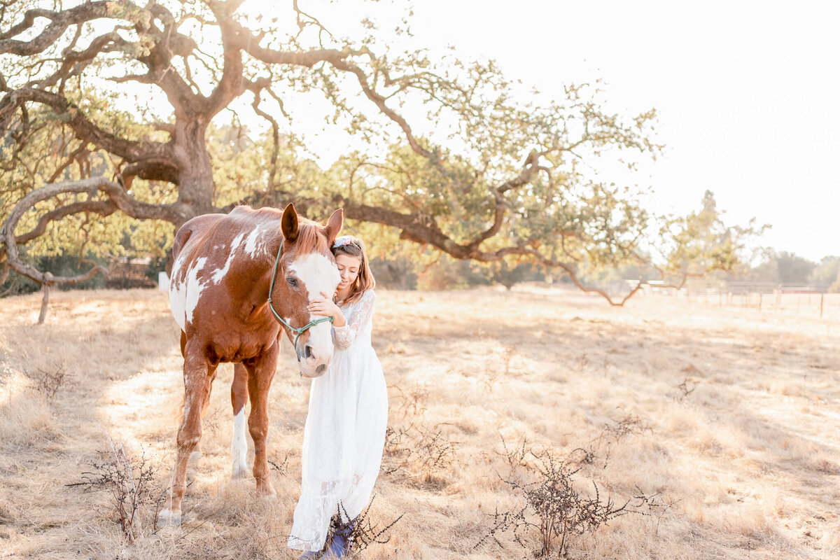 a girl in a long white dress standing in a field with her horse and snuggling him