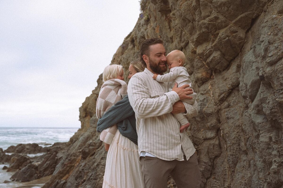 mother and father holding two babies at crescent bay beach near rock background