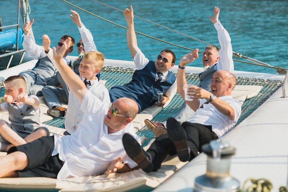 Male guests relax on a catamaran waving at other other guests while cruising the coastline