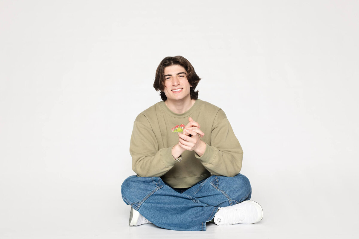 A young man with medium-length dark hair is sitting cross-legged on the floor against a plain white background. He is wearing a beige sweater, blue jeans, and white sneakers, and he is smiling while holding his hands together.