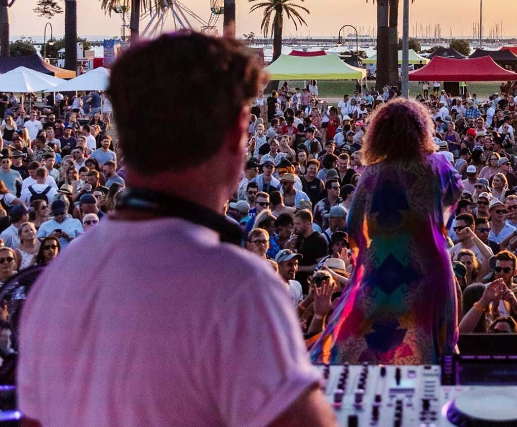 Backlit view of the crowd and performers with colourful tents and palm trees at sunset during St Kilda Festival