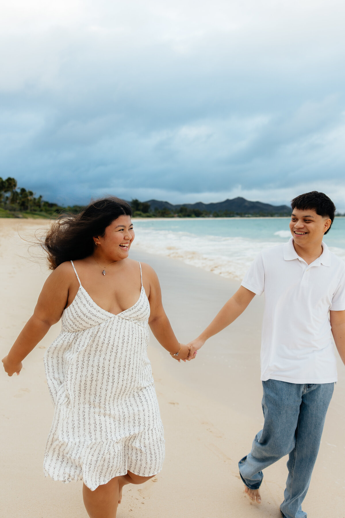 Couple walking hand in hand along the beach in Oʻahu, smiling and laughing together during their photoshoot at Kailua Beach