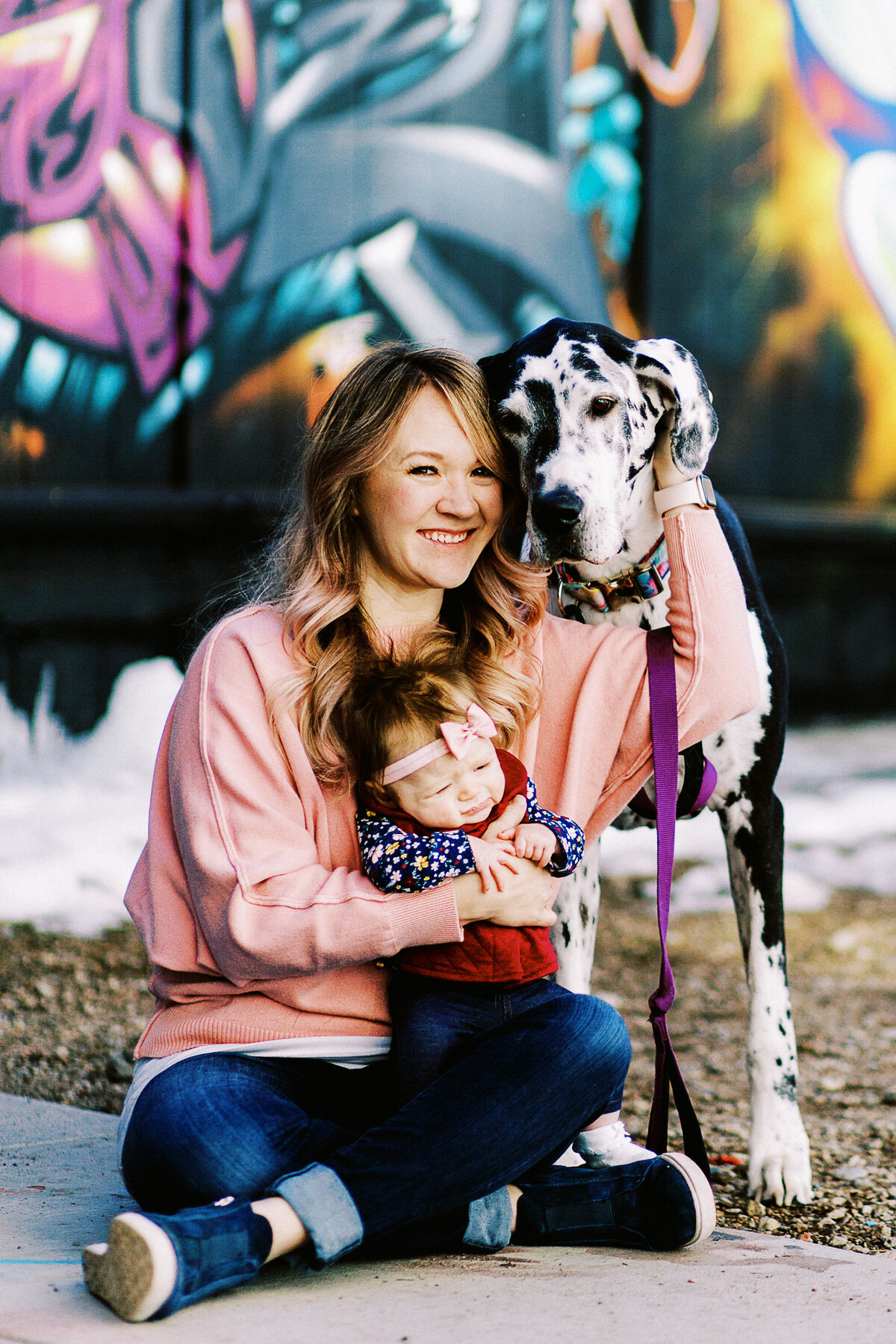 a mom in pink holds her baby girl and tall great dane while sitting on urban sidewalkd