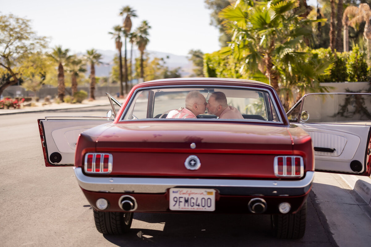 Rear view of red vintage Mustang with couple seated inside, driving into the golden Palm Springs landscape.