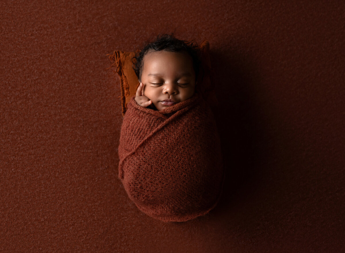 Baby boy wrapped in rust-colored knit with matching backdrop, hand posed gently by cheek | Newborn studio session near Lakeway and Austin