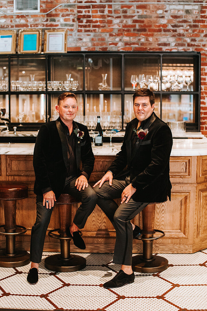 Two grooms wearing black tuxedos sitting on bar stools in front of a wooden bar.