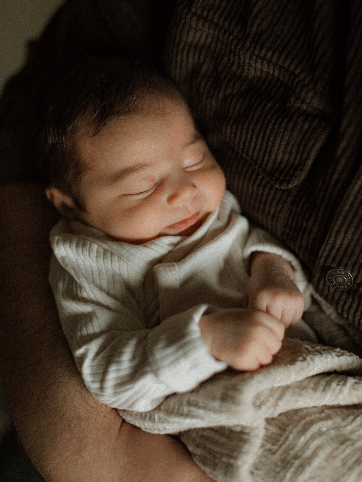 A close-up of a peacefully sleeping baby wrapped in a cream-colored blanket, nestled in dad’s arms.