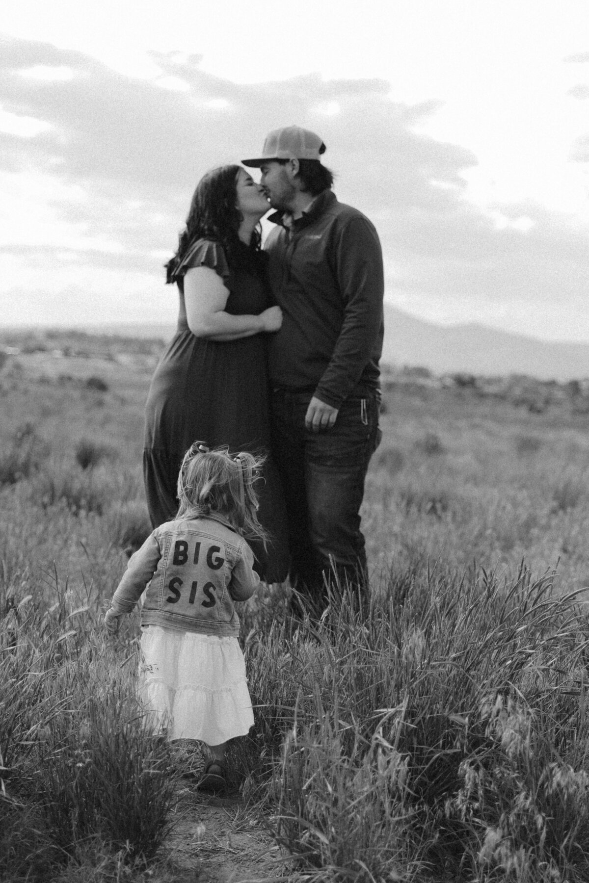 Candid Family Maternity Photo of Couple Kissing with Daughter Wearing “Big Sis” Jacket in Field