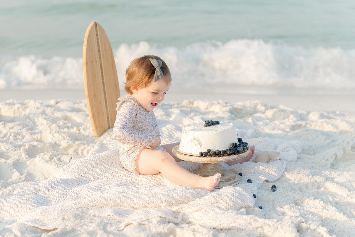 A baby girl is sitting on the beach with a surfboard in front of her smash cake captured by a professional destin family photographer.