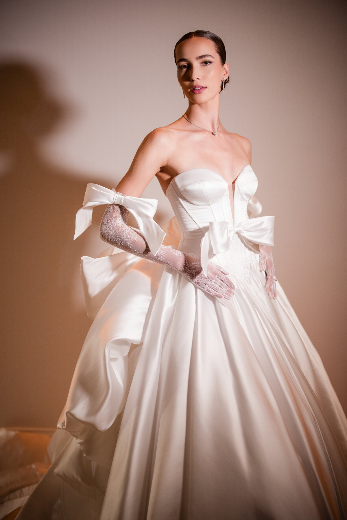 A bride wearing an elegant white wedding dress with bow details and lace gloves stands against a softly lit background. This photo was taken by a destination wedding photographer for European castle weddings