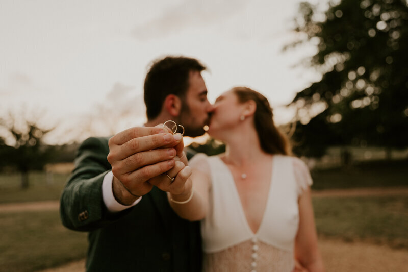 Mariés flous en arrière plan échangeant un baiser dans un cadre extérieur et montrant leurs alliances nettes au premier plan. Photo prise par Laura Termeau photographie.