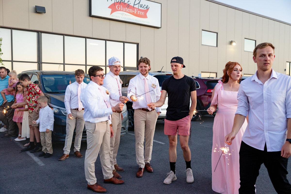 A group of wedding guests, including groomsmen in white shirts and pink ties, hold sparklers and smile as they celebrate outside the venue during the evening.