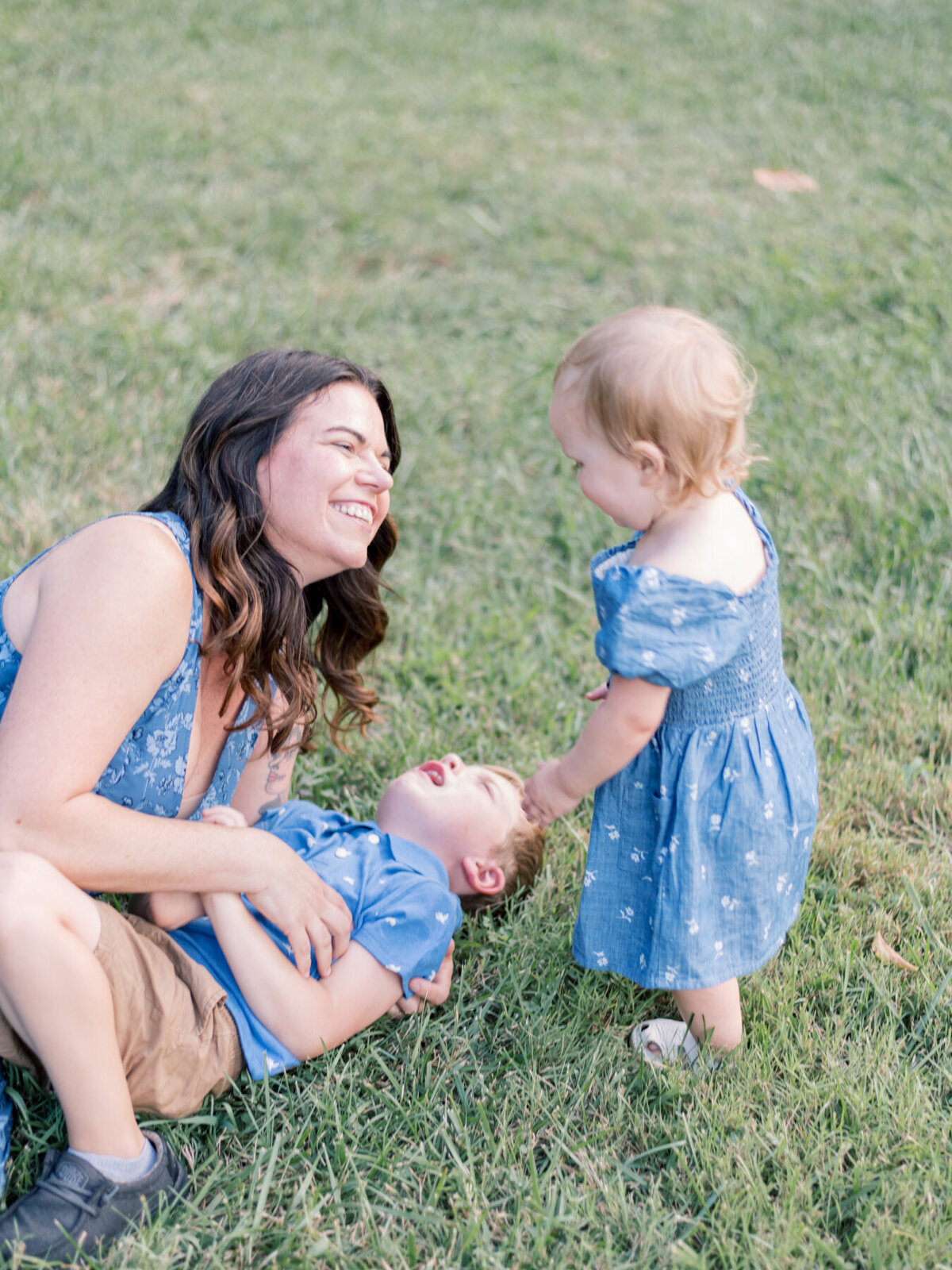 Young daughter laughing at Mom and brother who are wrestling on grass.