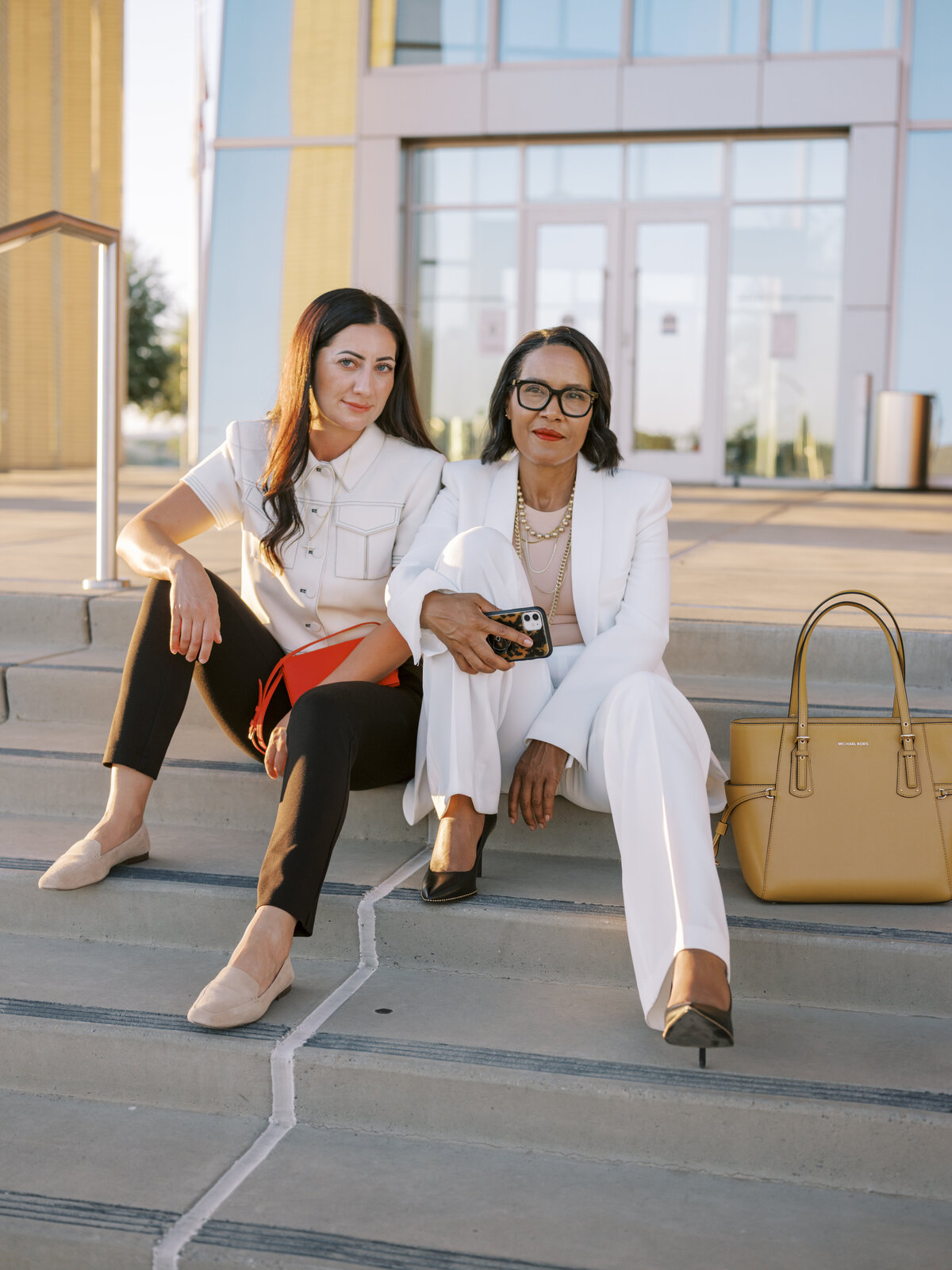 Two stylish women seated on outdoor steps in white and black suits during NYC brand photo session.