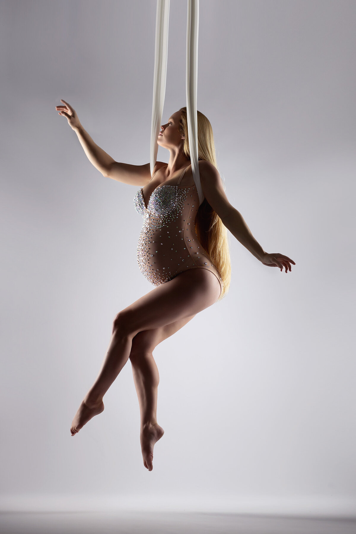 Woman gracefully suspended from white aerial silks, wearing a sparkling rhinestone-studded bodysuit. Her long blonde hair flows down as she reaches out, creating a stunning pose against the minimalist backdrop. DFW maternity photographer.