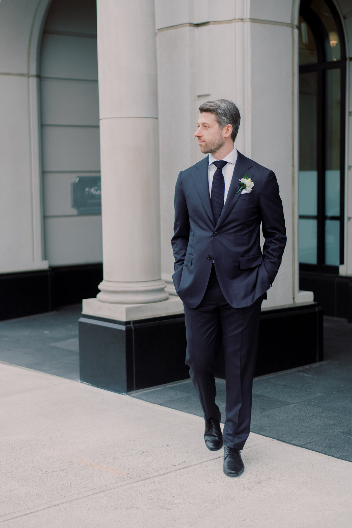 A full-body portrait of a groom walking confidently outside a classic architectural setting, wearing a navy suit with a white flower boutonniere